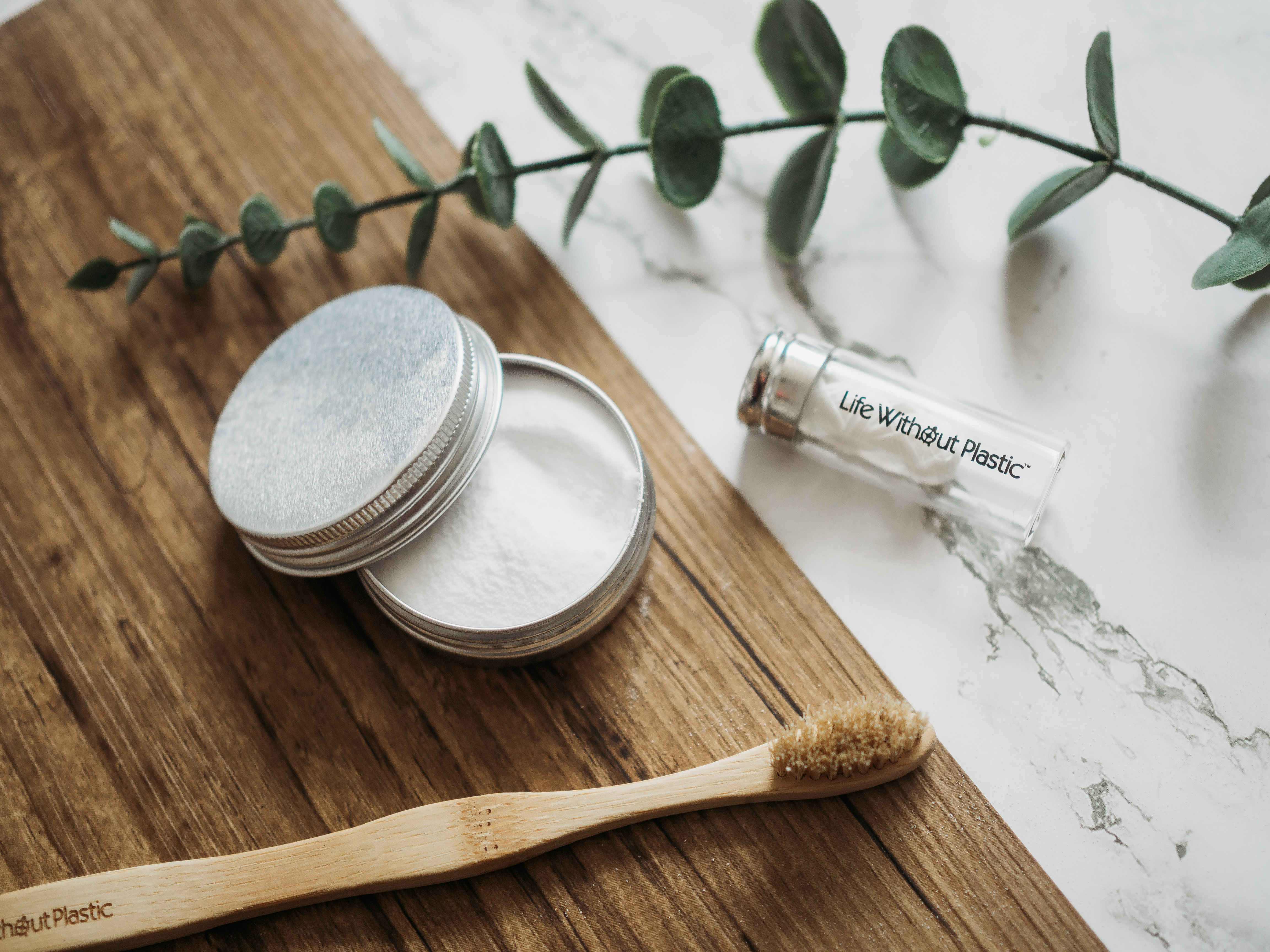 Life Without Plastic's compostable toothbrush, silk floss in a reusable glass jar, and baking soda in tin container on a wooden platform.